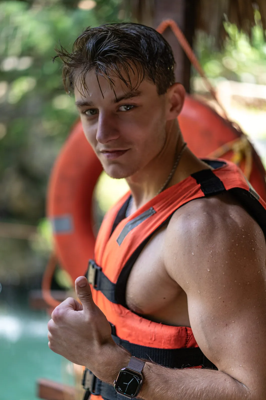 A fit young man wearing an orange life vest, giving a thumbs-up with confidence near a cenote