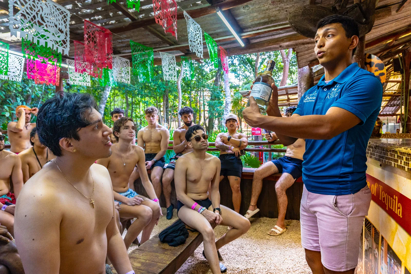 A group of shirtless men attentively listening to a guide presenting a bottle, surrounded by colorful decorations in an open-air setting
