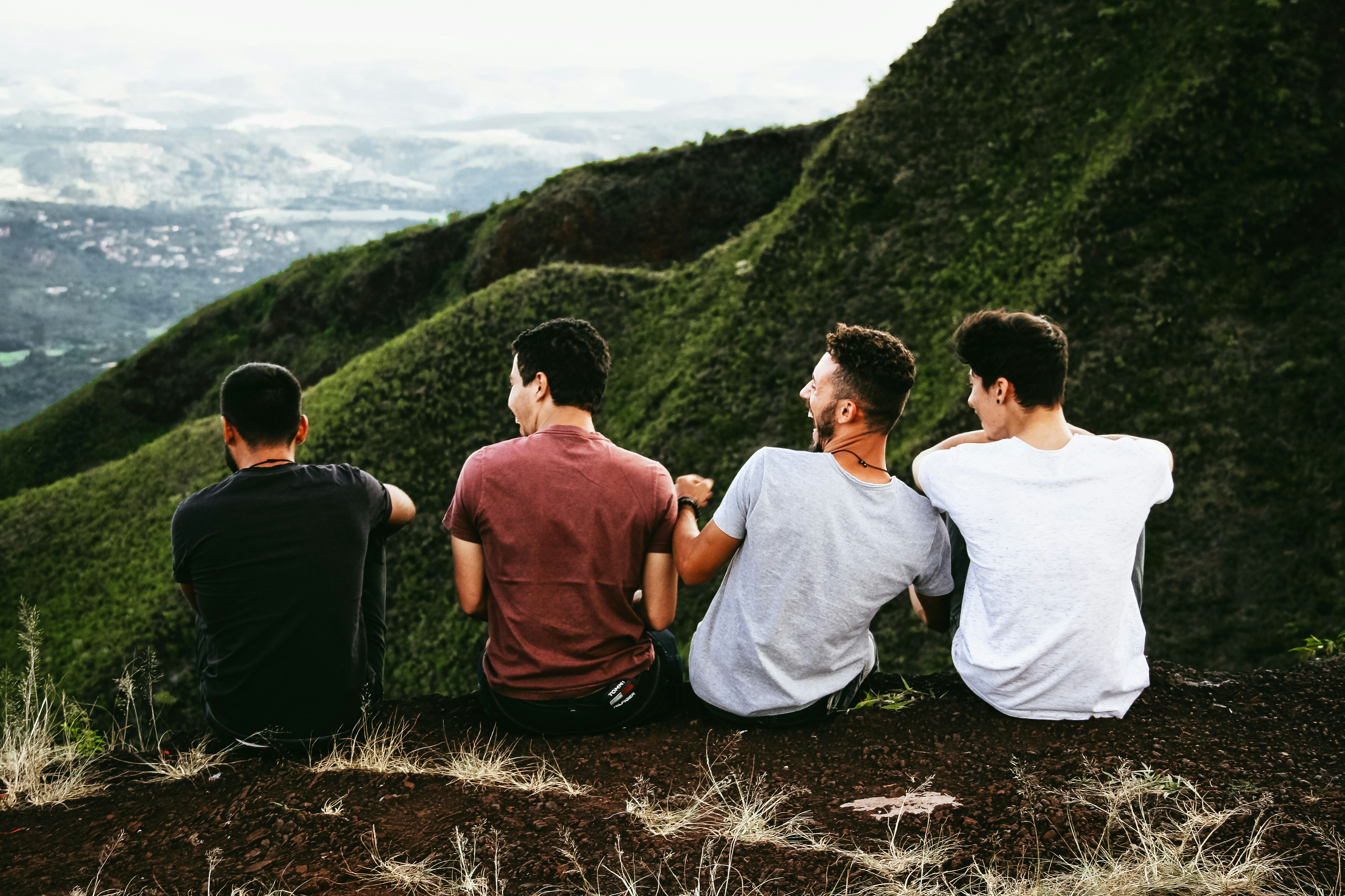 Four men sit side by side on a hilltop overlooking green mountains and a distant valley