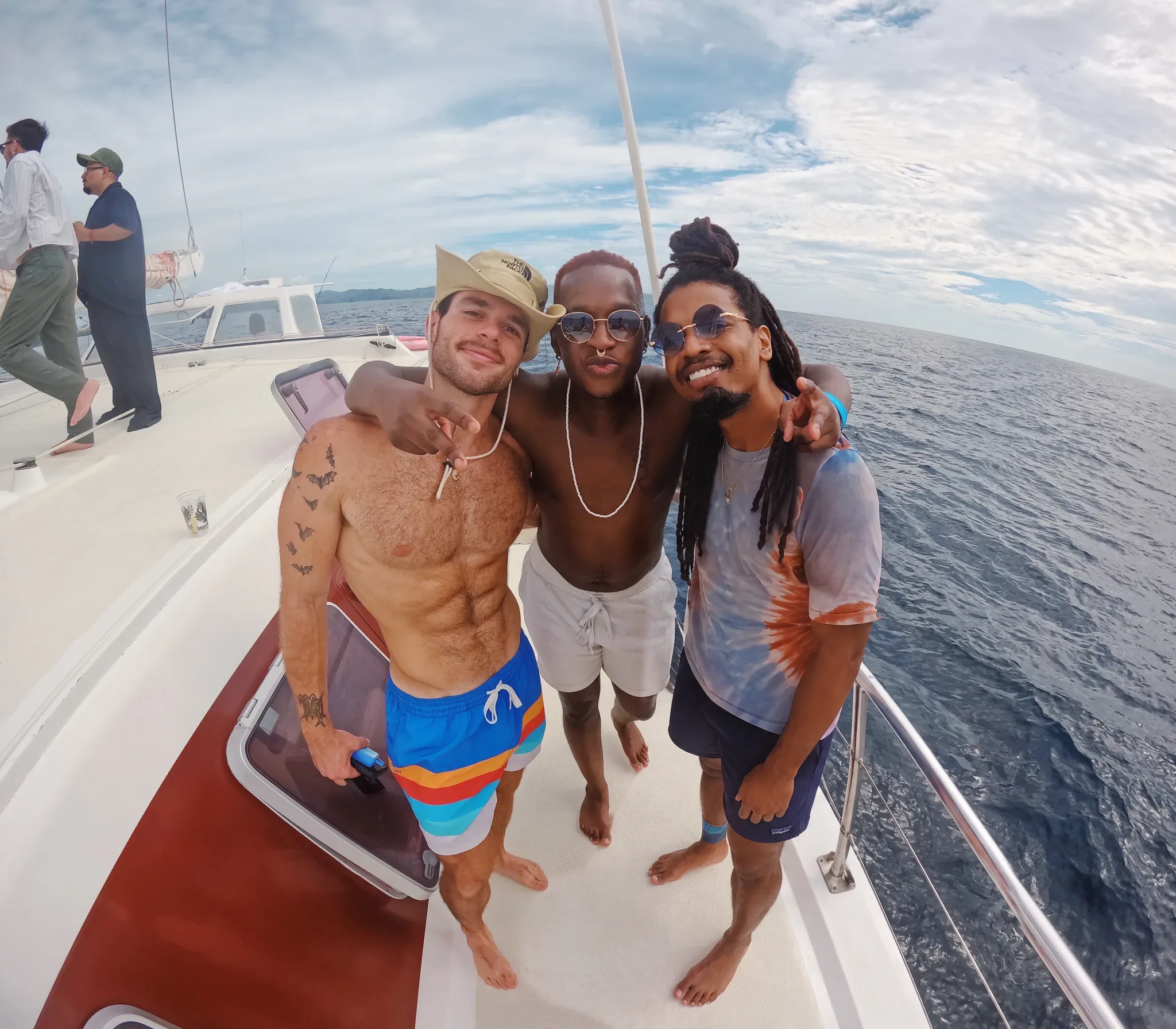 Three men smiling together on the deck of a catamaran on a Visiting Wrld retreat yacht day, open sea behind them