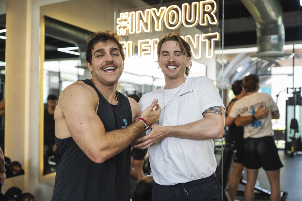 Two men smiling at the camera embracing each other with a neon sign in the back