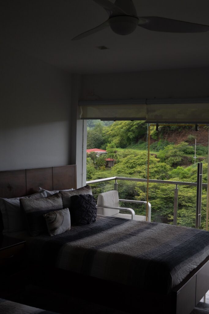 A dimmed bedroom with a Queen sized bed and a balcony overlooking trees and greenery.